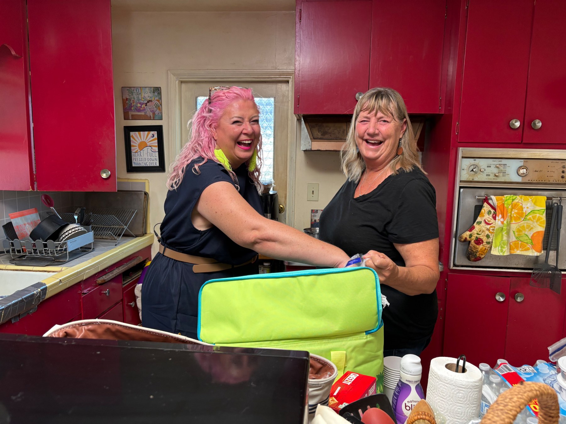 Host Unison Frances, with pink hair, laughing with a woman in a kitchen with bright red cabinets.