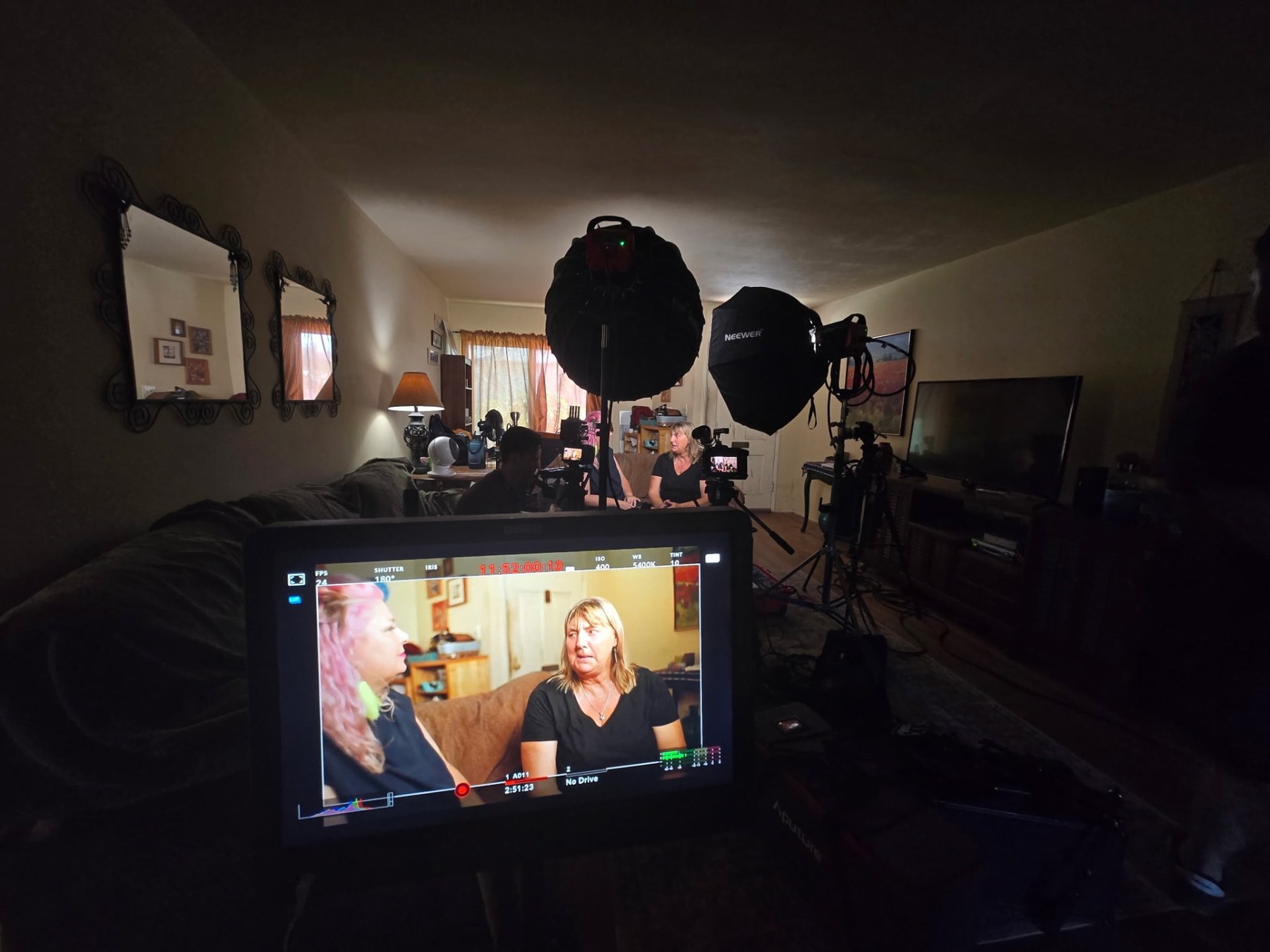 A camera monitor in the foreground shows two women being interviewed on a couch, with lighting and crew around the room.