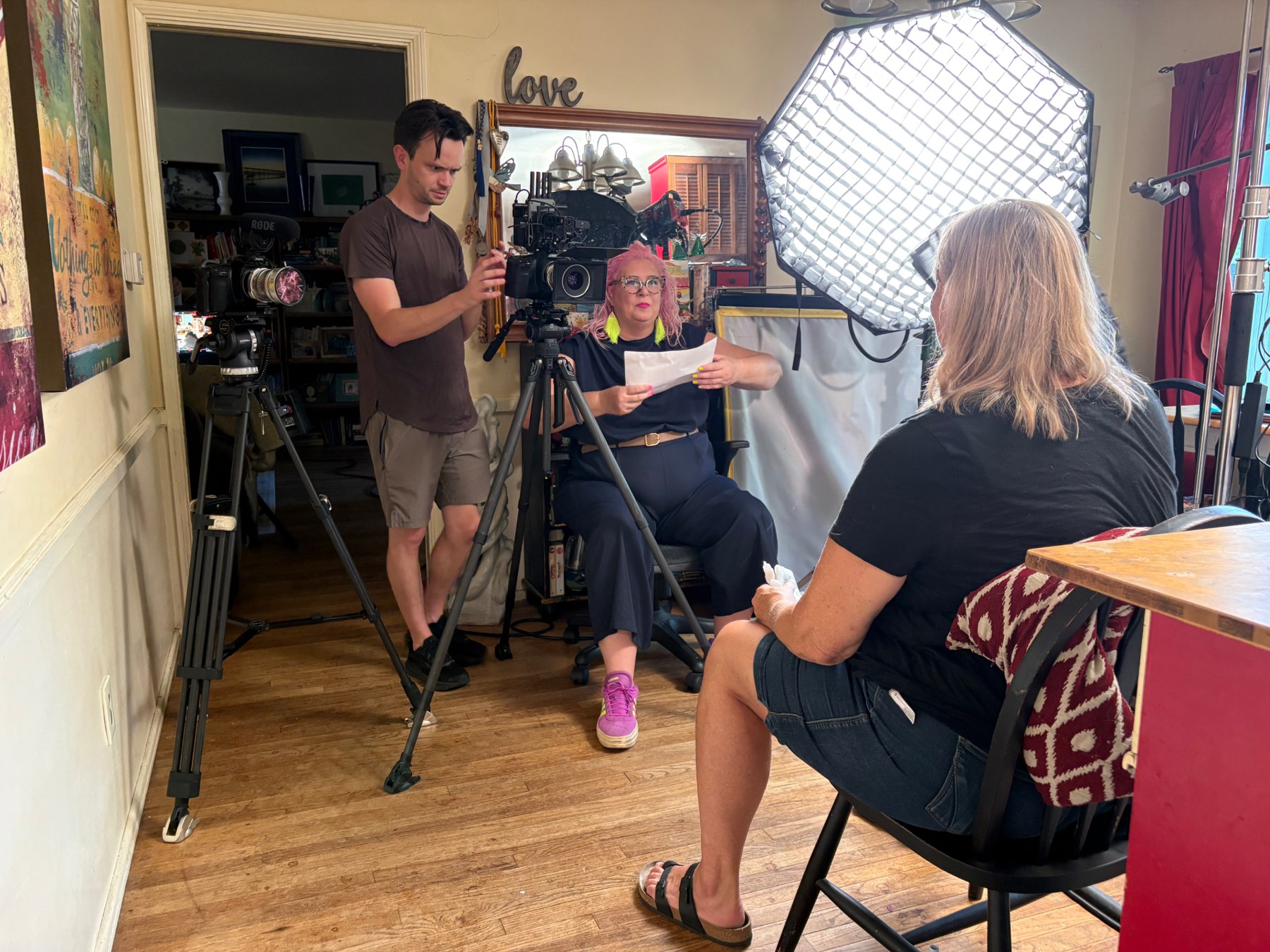 Host Unison Frances, with pink hair, reads from a page during an interview in a living room while a camera operator films.